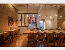 Ein gemütlicher Speiseraum mit Backstein- und weißen Wänden, ausgestattet mit langen Holztischen, gedeckten Plätzen und hölzernen Stühlen. Im Hintergrund ist eine offene Küche mit modernen Geräten sichtbar, daneben ein Kühlschrank mit Getränken; dekorative Hänge- und Wandlampen erhellen den Raum.