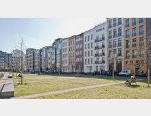 Reihen moderner Wohngebäude in unterschiedlichen Farben und Fassadenmaterialien säumen eine offene Grünfläche mit Sitzbänken und gepflasterten Gehwegen. Im Vordergrund sind kahle Bäume und parkende Autos zu sehen, während Personen auf den Bänken sitzen.