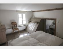 Ein Schlafzimmer mit niedriger Decke, weißen Wänden und Holzboden. Im Raum stehen ein Doppelbett mit weißer Bettwäsche, ein Holzstuhl, eine kleine Kommode und ein Fenster mit Tageslicht; ein Durchgang führt zu einem angrenzenden Raum mit kariertem Boden.