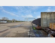 Industriegelände mit betonierter Fläche, einem großen Kohlehaufen auf der rechten Seite und einer Kiesförderanlage im Hintergrund. Links am Flussufer liegt ein Frachtschiff festgemacht, im Hintergrund Bäume und ein blau bewölkter Himmel.