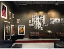 Galerieraum mit moderner Einrichtung, an einer anthrazitfarbenen Wand hängen großformatige Farbfotografien von Blumen und Schwarz-Weiß-Bilder von Models in verschiedenen Posen. Im Vordergrund stehen Ledersessel, ein schlichter Tisch mit einer Designlampe und einige schwarze Sitzmöbel.
