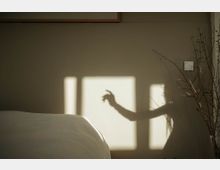 Ein minimalistisches Schlafzimmer mit beiger Wand, auf der Licht und Schatten eines Fensters sowie der Silhouette einer Hand sichtbar sind. Im Vordergrund ist ein Teil eines Bettes zu sehen, rechts stehen dünne Zweige in einer Vase.