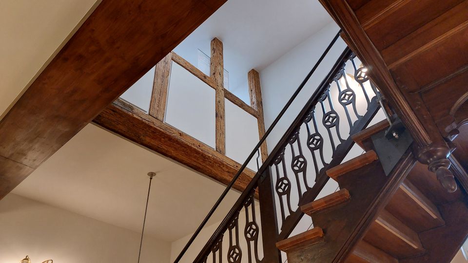 Innenansicht eines Treppenhauses mit einer Holztreppe, einem verzierten Geländer aus dunklem Metall und sichtbaren Holzbalken im oberen Bereich der Wand. Die Wände sind weiß gestrichen, und eine einzelne Pendelleuchte hängt von der Decke.