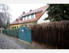 Straßenszene mit einem Haus im Landhausstil, gelber Fassade und rotem Ziegeldach; daneben ein Holzzaun mit einer grünen Metalltoröffnung. Die Straße ist mit Kopfsteinpflaster belegt und von Bäumen gesäumt.