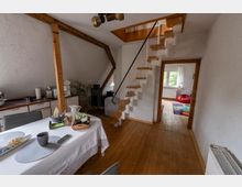 Offener Wohn- und Essbereich mit Holzdielenboden, weiß verputzten Wänden und sichtbaren Holzbalken; links eine Küchenzeile und ein Esstisch. In der Mitte führt eine schmale Holztreppe zu einer Dachluke, rechts ein Durchgang in ein Zimmer mit Fenster und kleinem Teppich.