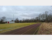 Leichtathletikanlage im Freien mit Laufbahn entlang einer großen Rasenfläche, daneben ein kleiner Funktionsbau mit rotem Tor und ein hoher Flutlichtmast. Im Hintergrund stehen ein überdachter Unterstand und eine Baumreihe unter bedecktem Himmel.