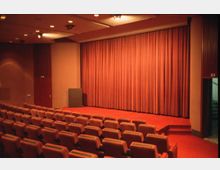 Kleiner Vortragssaal mit roten gepolsterten Sitzreihen, einem roten Vorhang auf der Bühne und warm beleuchteter Deckenbeleuchtung. Auf der rechten Seite führt eine Treppe aus dem Raum.