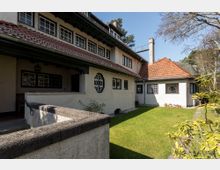 Wohnhaus im traditionellen Stil mit heller Putzfassade, roten Ziegeldächern und mehreren Fenstern mit Sprossenglas. Im Vordergrund befindet sich ein gepflegter Garten mit Rasen und umliegenden Bäumen.
