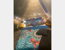 Gemütliches Schlafzimmer in einer Dachschräge mit unverputzter Backsteinwand. Auf dem Bett liegt bunte Bettwäsche mit floralen Mustern und Vogelmotiven, eingerahmt von zwei kleinen Nachttischen mit Lampen.