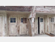 Eine Reihe alter, verwitterter Holztüren in einem überdachten Eingangsbereich, erkennbar an abblätternder Farbe und kleinen Fenstern mit Sprossen. Holzbalken stützen das Vordach, und die Struktur zeigt deutliche Spuren von Verwitterung.