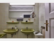 Ein Badezimmer mit zwei grünen Waschbecken, pastellfarbigen Fliesen mit floralen Mustern, einem großen Spiegel und zwei grünen Ablageflächen. Ein Handtuchhalter mit aufgerollten Handtüchern und diverse Gegenstände wie ein Seifenspender und Dekorationen befinden sich im Raum.