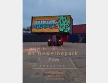 Ein Industriehof mit einem gelben Schiffscontainer, der in der Luft schwebt und mit bunten Graffitis bemalt ist. Im Hintergrund sind weitere Container sowie eine graue, wolkige Himmelskulisse zu sehen. Text auf dem Bild verweist auf den „B1 Gewerbepark Film Location“ mit einer Adresse in Müncheberg.
