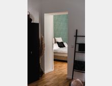 Ein Blick durch eine offene Tür in ein Schlafzimmer mit einem Bett, das weiße Bettwäsche und schwarze Kissen hat; die Wand ist mit grünem Muster tapeziert. Im Vordergrund sind ein schwarzer Schrank, eine Glasvase mit Pampasgras und eine schwarze Leiter-Dekoration zu sehen. Der Boden besteht aus Holz.