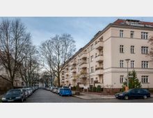 Wohnstraße mit einem beige verputzten Mehrfamilienhaus im Vordergrund, das mehrere Balkone aufweist. Die Straße ist mit Kopfsteinpflaster bedeckt und gesäumt von parkenden Autos sowie kahlen Bäumen.