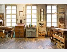 Eine traditionelle Werkstatt mit hölzerner Einrichtung, darunter breite Werkbänke, ein Schrank mit Schubladen und Werkzeugaufbewahrung. An der Rückwand befinden sich drei große, alte Fenster, sowie diverse Werkzeuge, Holzstücke und Zeichnungen an der Wand.