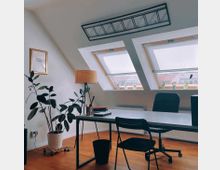 Ein modernes, minimalistisches Arbeitszimmer unter einer Dachschräge mit zwei großen Dachfenstern, durch die Tageslicht einfällt. Der Raum enthält einen Schreibtisch mit Stuhl, eine stehende Zimmerpflanze, eine Stehlampe, sowie kleine Pflanzen und Dekorationen auf der Fensterbank.