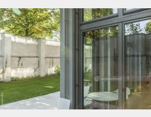 Moderner Außenbereich mit Rasenfläche und heller Steinterrasse neben einer Glasfassade. Im Hintergrund eine hohe, cremefarbene Mauer mit Kletterpflanzen, dahinter große Bäume.