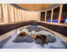 Innenraum eines Schlafbereichs in einem Fahrzeug mit abgerundeten Wänden, gepolsterten Seitenlehnen und großen Fenstern. Zwei Personen liegen auf einem Bett mit blauen Decken und nautischen Motiven, während die Vorhänge teils geöffnet sind und bunte Lichter draußen sichtbar sind.