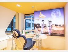 Heller, moderner Behandlungsraum in einer Zahnarztpraxis mit gelber Decke, dunklem Behandlungsstuhl, zahnärztlichen Geräten und einer großen Wandgrafik mit Astronauten auf einer Mondlandschaft im Hintergrund.