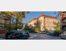 Ecke einer ruhigen Wohnstraße mit gepflastertem Straßenbelag, geparkten Autos und von Bäumen gesäumten Gehwegen. Im Hintergrund ist ein mehrstöckiges Gebäude im historischen Stil mit roten Dächern und hellen Fassaden sowie Balkonen zu sehen.