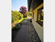 Gepflasterter Weg entlang eines beige gestrichenen Hauses mit angrenzendem Kiesbeet und Fenstern. Rechts des Wegs sind blühende Pflanzen und ein roter Baum, im Hintergrund Hecken und ein Rasenbereich unter klarem Himmel sichtbar.