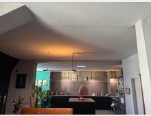 Moderne Wohnküche mit dunklen Schränken, zentraler Kochinsel und grauer Wandverkleidung. Im Hintergrund sind Pflanzen und Dekoelemente zu sehen, während die Deckenbeleuchtung warmes Licht abstrahlt.