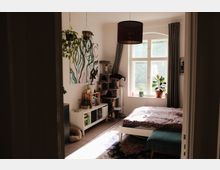 Helles Schlafzimmer mit weißem Bett auf gemustertem Teppich, großer Bogenfensterfront mit dunklen Vorhängen und mehreren Zimmerpflanzen auf der Fensterbank. An der Wand hängen ein abstraktes Bild und Hängepflanzen; daneben stehen ein Katzenkratzbaum und ein niedriges Regal mit Büchern.