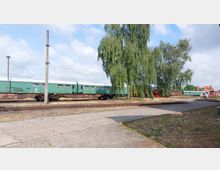 Ein Bahnhofsgelände mit einer grünen, abgestellten Personenbahn auf Schienen, daneben ein großer Baum. Im Hintergrund sind weitere Schienen, historische Lokomotiven und ein Gebäude mit rotem Dach zu sehen.