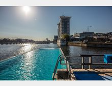 Ein schwimmender Pool am Ufer eines Kanals mit Blick auf ein hohes, modernes Gebäude und eine Skulptur im Wasser; im Hintergrund sind weitere Gebäude und Stege sichtbar bei sonnigem Wetter.