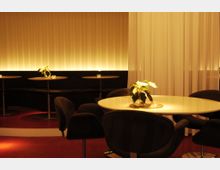 Ein stilvoll eingerichteter Innenraum eines Cafés oder einer Lounge mit runden Tischen, gepolsterten Stühlen und indirekter Beleuchtung an einer gerillten Wand. Auf den Tischen stehen kleine Pflanzen und Teelichter als Dekoration.