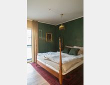 Schlafzimmer mit grünen Wänden, einem Holzbett mit vier Pfosten und heller Bettwäsche. Der Raum hat einen roten Teppich, einen Kronleuchter, ein Fenster mit cremefarbenen Vorhängen und eine gerahmte Wanddekoration.