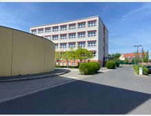 Ein moderner, vierstöckiger Gebäude mit weißen Wänden und roten Fensterrahmen steht in einem gepflasterten Bereich, umgeben von niedrigen Bäumen und Büschen. Links im Vordergrund ist eine gelbe, abgerundete Wand zu sehen, rechts sind Parkplätze und einzelne Laternen erkennbar.