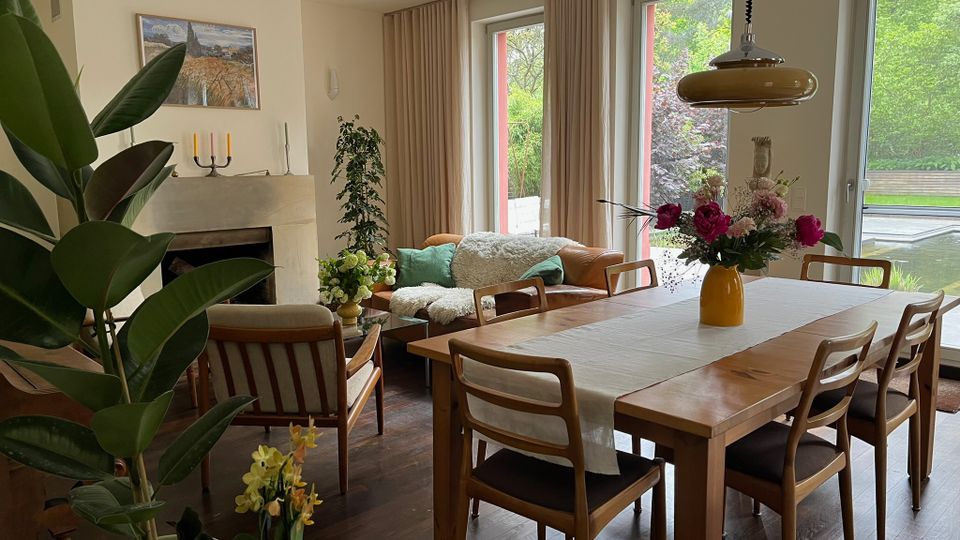 Heller Wohn- und Essbereich mit Holzesstisch und Stühlen, Ledersofa neben einem Kamin sowie mehreren Zimmerpflanzen. Große bodentiefe Fenster mit Vorhängen öffnen den Blick ins Grüne und auf eine Terrasse mit Wasserfläche.