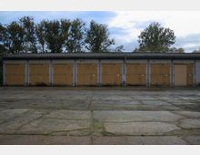 Einstöckige Garagenreihe mit gelblichen Holztoren und einer Fassade aus hellen Ziegeln, davor eine große, mit Betonplatten gepflasterte Fläche. Im Hintergrund sind Bäume und ein wolkiger Himmel zu sehen.