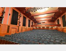 Ein Innenraum eines Theaters mit mehreren Sitzreihen aus blauen Polstersesseln, holzvertäfelten Wänden und einer Empore. An den Wänden befinden sich große schwarze Vorhänge, und an der Decke hängen Beleuchtungseinrichtungen.