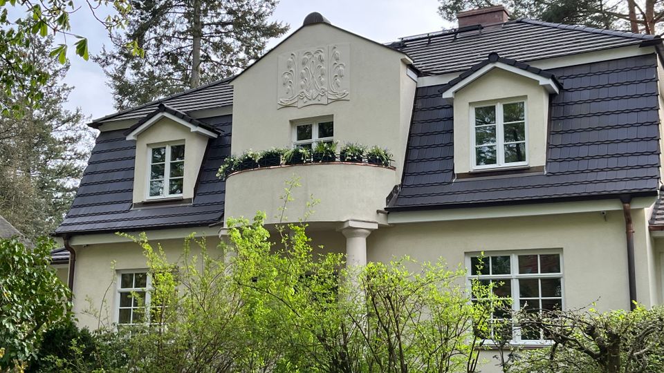 Zweistöckiges Wohnhaus mit cremefarbener Fassade und dunkel gedecktem Mansarddach, mit zwei Gaubenfenstern und einem mittigen, halbrunden Balkon mit Blumenkasten. Im Vordergrund stehen dichte Sträucher und ein Zaun, das Haus liegt in einer von hohen Bäumen umgebenen Garten- oder Waldumgebung.