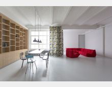 Moderner, heller Raum mit weißem Boden und gewölbter Decke; links eine Holzregalwand mit integriertem Sideboard, in der Mitte ein großer Tisch mit Stühlen und Hängelampen, rechts ein leuchtend rotes Sofa und ein Vorhang mit buntem Muster vor einem großen Fenster.