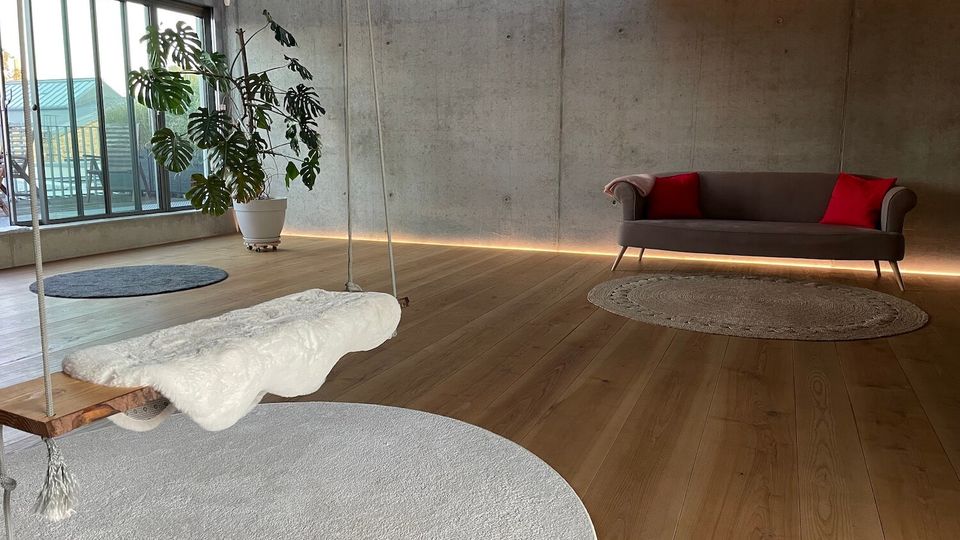 Minimalist loft-style living room with concrete walls, wide wooden floors, and large industrial windows overlooking a balcony. A rope-hung wooden swing with a white cushion sits over a round rug, with a potted monstera plant and a gray sofa with red pillows on the far wall.