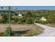 Wegweiser mit der Aufschrift „Panoramaweg Werderobst“ in einer ländlichen Umgebung mit Feldern, Bäumen und vereinzelten Häusern. Im Hintergrund sind ein See, Wald und eine kleine Stadt auf einem Hügel sichtbar.