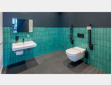 Barrierefreies Badezimmer mit grünen Fliesen an der Wand, schwarzem Boden, einem wandhängenden Waschbecken mit Spiegel und einem wandmontierten WC mit seitlichen Haltegriffen.