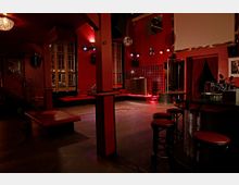 Ein dunkler Raum in einem Nachtclub mit roten Wänden und Sitzbänken. Im Vordergrund stehen Barhocker mit roten Polstern, im Hintergrund ein Podium, Glasbaustein-Elemente und ein Loungebereich mit gedämpfter Beleuchtung.