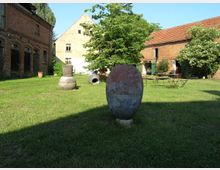 Ein Innenhof mit einer Rasenfläche, umgeben von Backsteingebäuden mit roten Ziegeldächern und grünen Fensterläden. Im Vordergrund stehen mehrere große, runde Steingefäße, während im Hintergrund ein Baum und Gartenmöbel zu sehen sind.