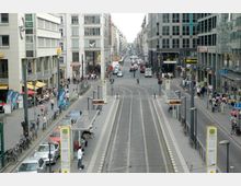 Straßenszene in einer belebten Innenstadt mit Straßenbahngleisen in der Mitte, flankiert von Haltestellen mit Fahrgastunterständen. Umgeben von mehrstöckigen Büro- und Geschäftsgebäuden mit Schaufenstern, Straßencafés und Fußgängern auf den Gehwegen.