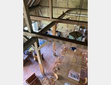 Ansicht eines offenen Raumes mit rustikalem Holzgebälk und Ziegelfußboden. In der Mitte stehen große Holztische, umgeben von Holzstühlen, während bunte Wimpelketten von den Balken hängen.