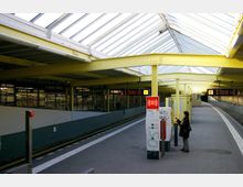 Bahnhofshalle einer erhöhten Stadtbahnstation mit gelben Stahlträgern und einem Dach aus Glasplatten. Im Vordergrund befinden sich ein SOS-Notrufterminal und ein Fahrgast, im Hintergrund verlaufen die Gleise neben den Bahnsteigen mit Anzeigetafeln und einer Uhr.