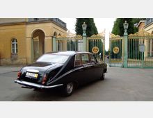 Ein schwarzes Oldtimer-Auto steht vor einem großen, kunstvoll verzierten Tor mit goldenen Ornamenten und grünen Rahmen. Im Hintergrund sind symmetrisch angeordnete Bäume und gelbe Gebäude mit Rundbögen zu sehen.