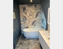 Modernes Badezimmer mit freistehender weißer Badewanne vor einer großen Wandfläche mit Koi-Fisch-Motivtapete. Dunkle Wände, graue Steinfliesen am Boden und ein weißer Waschtisch neben einem beleuchteten Spiegelrahmen prägen den Raum.