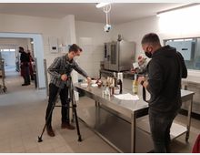 Moderne Großküche mit Edelstahl-Arbeitsflächen und Fliesenboden. Zwei Personen mit Kameras und Masken arrangieren Produkte auf einem Tisch, während im Hintergrund mehrere Personen in Arbeitskleidung zu sehen sind.