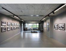 Ein moderner Ausstellungsraum mit grauen Wänden, an denen schwarz-weiße Fotografien hängen, und einer Betondecke mit Deckenspots. Im Hintergrund ein verglaster Bereich mit Möbeln und Blick ins Grüne.