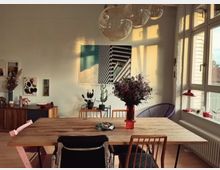 Esszimmer in einer Wohnung mit einem Holztisch und verschiedenen Stühlen, dekoriert mit Blumen in einer roten Vase. Im Raum hängen Gemälde an den Wänden, und große Fenster lassen Tageslicht einfallen, während eine moderne Hängeleuchte an der Decke hängt.